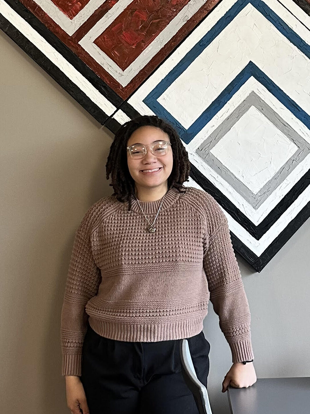 Daiah stands in front of a large painting of the Kennari logo. She is wearing a light brown sweater, black pants, and is smiling.