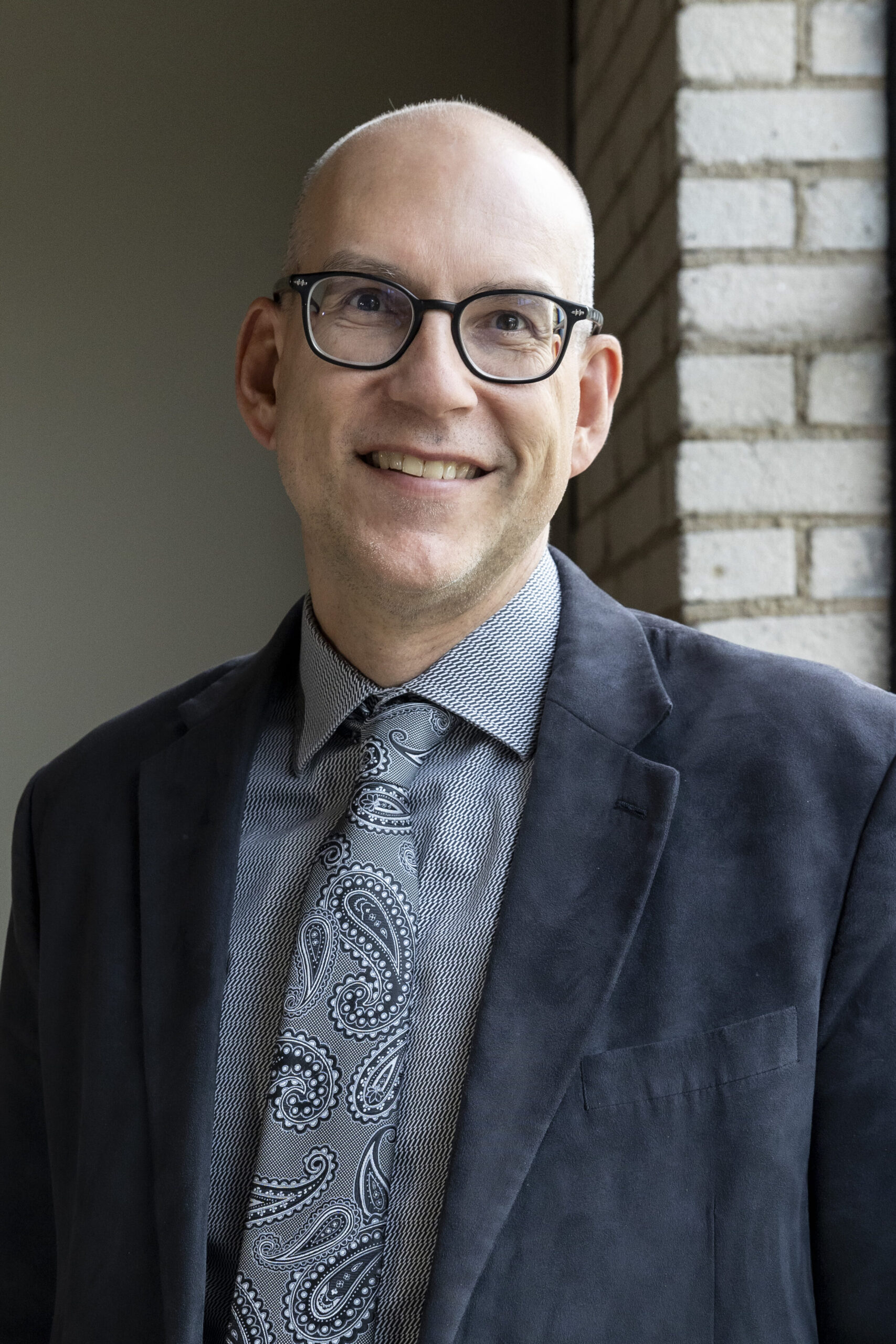 A headshot of Andy DeBraber. Andy is smiling, with semi-round black glasses, a navy suit, and light blue patterned dress shirt with paisley tie.